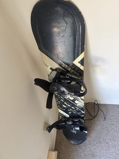 A black and white snowboard with black bindings is propped vertically against a white wall, leaning slightly. The snowboard has a distressed graphic design with a marble-like pattern in black and white, and a band of text that appears to be repeated "MISSION". A black cable is coiled on the carpeted floor next to the snowboard. The environment is an indoor space with a light-colored wall and carpet. The image appears to be taken in the City of Binghamton, United States of America.