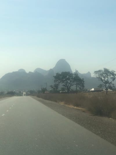 The scene depicts a rural road in Dott, Nigeria, extending forward into a hazy landscape under a pale blue-grey sky. The asphalt road features white dashed center lines and a gravel shoulder on the right. Both sides of the road are lined with dry, brownish vegetation and scattered deciduous trees with sparse foliage. Utility poles with overhead wires run parallel to the road on the right. In the mid-ground on the right, two small, single-story structures with light-colored roofs are partially visible behind the vegetation. In the distant mid-ground, a faint silhouette of a vehicle is discernible on the road, and a cluster of buildings indicative of a small settlement is present on the left. The background is dominated by a range of prominent hills and mountains, appearing as hazy, blue-grey silhouettes with rounded and peaked formations. No individuals are clearly visible.