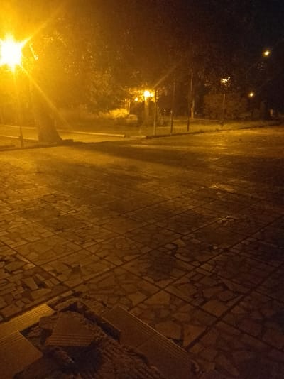The image depicts a nighttime scene on a paved surface in Gorno Aleksandrovo, Bulgaria. A bright street lamp illuminates the area from the left, casting a warm orange glow and creating strong shadows. Trees and other foliage are visible in the background, also lit by the lamp. The ground is covered in square tiles, some of which appear wet, reflecting the light. In the foreground, there are some broken tiles and debris, suggesting a state of disrepair or ongoing work. The overall atmosphere is dark and somewhat eerie due to the low light conditions and the illumination from the single light source. There are no people or animals visible in the frame.