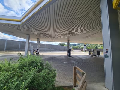 The image captures a view of a gas station canopy in Sankt Marein im Mürztal, Austria. The canopy has a ribbed, light-colored ceiling supported by grey columns. A yellow pipe runs along the edge of the canopy. The ground beneath the canopy is paved with interlocking grey bricks. Several gas pumps are visible, one of which is a black and white dispenser with a red fire extinguisher attached. To the left of the scene, a dense green bush grows. In the background, a tall grey sound barrier wall runs along a road, with trees and hills visible beyond it. The sky is partly cloudy. There are no people visible in the image.