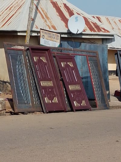 The image displays a street scene in Bahit, Nigeria, featuring multiple metal doors and gate sections leaning against a building. In the foreground, two maroon-colored metal doors, each with gold-colored decorative inserts, are positioned upright on a paved curb. To their left, a dark-colored metal gate section featuring a diagonal slatted pattern is visible, along with additional dark metal frameworks. The background reveals a light-colored building with a weathered, rusted corrugated metal roof. A white satellite dish is mounted on the roof. A sign board, reading "THE REDEEMED CHRISTIAN CHURCH OF GOD - LIVING GRACE ASSEMBLY" and listing service times, is affixed to the building wall above the metal items. The immediate foreground shows an asphalt road. No individuals are present.
