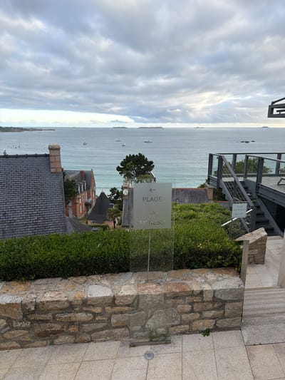 An overcast day in La Clarté, France, offers a view of the sea with several boats dotted on the water. In the foreground, a glass sign with an arrow pointing left and the word "PLAGE" (beach) is positioned in front of a stone wall and lush green bushes. To the right of the sign, another smaller sign indicates "VILLA EDEN ROC" with arrows and numbers. A modern metal staircase with glass railings leads up to a deck area on the right side of the frame. Behind the bushes, residential buildings with slate roofs and red-brick facades are visible, nestled amongst trees. The overall scene depicts a coastal landscape with a mix of natural beauty and modern architecture.