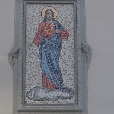 This is a mosaic artwork depicting Jesus Christ, likely the Sacred Heart of Jesus, in a horizontal orientation. He is portrayed with a halo and his right hand is placed over his chest, over a radiant heart symbolizing divine love. Jesus is wearing a red tunic and a blue cloak draped over his body. The background of the mosaic is a light-colored, speckled pattern. To the right of Jesus, there appears to be a cloud-like form rendered in shades of grey and white, suggesting a celestial or ethereal element.

The mosaic is framed by a decorative, light grey border with ornate, leaf-like embellishments at the top and bottom centers. The artwork is mounted on a plain, light-colored wall, which appears to be indoors given the lighting conditions. There is no discernible text within the image. The overall impression is one of religious devotion and artistic craftsmanship. The artwork's context in Calcinate, Italy, suggests it is likely part of a church or religious building, a common setting for such iconography.