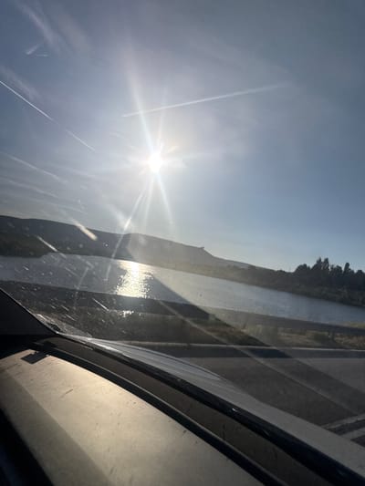The image captures a scenic view of a body of water, possibly a river or lake, reflecting the bright sun. The sun is prominently positioned in the sky, casting strong rays and creating a lens flare effect. In the foreground, the dashboard of a vehicle is visible, suggesting the photo was taken from inside a car. Beyond the water, a hilly landscape with trees and vegetation lines the horizon. The sky is clear and blue, with some faint contrails from aircraft visible. The overall scene depicts a tranquil, sunlit day, likely in a natural setting. The location is identified as Dibich, Bulgaria.
