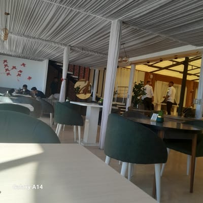 An indoor view of a restaurant in Khan Yunis, Unknown shows a draped ceiling with multiple white fabric panels. A few decorative red ribbons are pinned to a white wall on the left. Two men are seated at a table on the left, one looking at his phone and the other looking down at a white object, possibly a flower. Another man stands in the background, partially obscured. On the right, two men in white shirts and dark pants are standing near a table. Plants are visible in pots on some tables. The flooring appears to be tiled. In the foreground, a light-colored table is visible, with the text "Galaxy A14" overlaid in white.