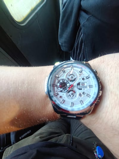 The image shows a close-up of a person's wrist wearing a white-faced chronograph watch with red accents. The watch has multiple sub-dials and a tachymeter scale on the bezel. The person is wearing a dark jacket over a striped shirt, and dark pants. The background appears to be the interior of a vehicle, possibly a bus or train, with a window visible on the left and dark seating. There is a keyring with a blue fob attached to the pants. The scene is not identifiable as being in the city of Sviatopetrivske, Ukraine, or any specific location.