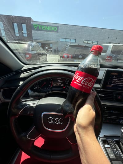 A first-person perspective shot from inside a car shows a hand holding a Coca-Cola bottle. The car's dashboard and steering wheel are visible. The background shows a building with a large "TENDO" sign, suggesting a commercial establishment. Several cars are parked outside, and a person is seen on a bicycle. The scene is set in Sarny, Ukraine.