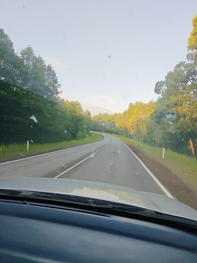 A road curves through a forest in Kamennyi Kliuch, Russia, with trees lining both sides of the asphalt. The view is from inside a vehicle, looking out through a windshield that shows some smudges and specks. The sky is a pale, hazy blue, with a hint of sunlight illuminating the tops of the trees on the right side of the road. The road itself appears to be in fair condition, with some cracks and tire marks visible. There are white markers on the side of the road to indicate the lanes. The overall scene suggests a peaceful drive through a natural landscape.