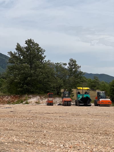 The image depicts a construction site in Jasenovo Polje, Montenegro. The foreground is a vast expanse of gravel. In the middle ground, there are three pieces of heavy machinery: two compactors and one asphalt paver. The compactor on the left is orange and black, the asphalt paver in the center is green and yellow, and the compactor on the right is orange with black accents. Behind the machinery, a dense, dark green forest with a large, mature tree dominates the scene. Further in the background, rolling hills covered in vegetation are visible under a cloudy sky. The overall impression is one of an active construction project in a natural, rural setting. There are no people visible in the image.