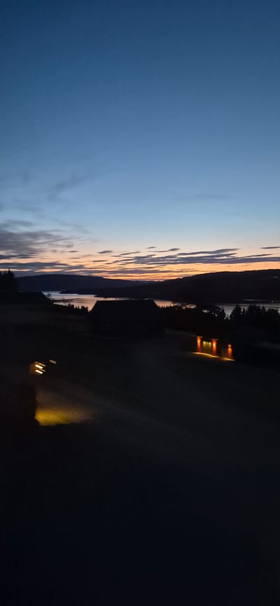 The image depicts a twilight scene over a body of water, likely Lake Mjøsa, near Biristrand, Norway. The sky is a gradient of deep blue at the top, transitioning to lighter blues and then to hues of orange and pink near the horizon, indicating the setting or rising sun. Wispy clouds are scattered across the sky.

In the foreground, the land is dark and silhouetted. Two dark structures, appearing to be buildings, are visible. One, closer to the center and slightly to the right, has several illuminated windows casting a warm, orange glow onto the surrounding dark ground. The other building is partially visible on the left, with a bright, yellowish light emanating from it, possibly from headlights or a floodlight, casting an elongated reflection on the ground. The ground itself appears to be a field or open area, also mostly in shadow.

In the distance, rolling hills or mountains form a dark silhouette against the brightly colored sky. The body of water stretches between these hills, reflecting the sky's colors and appearing as a band of lighter tones. Trees are sparsely scattered along the shoreline. The overall impression is one of a peaceful, serene evening in a rural or semi-rural setting.