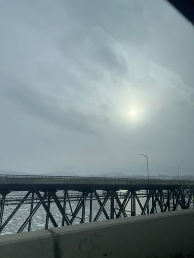 An overcast sky obscures the sun, casting a hazy glow over the scene. A bridge's steel framework spans over water, possibly a river. The water below reflects light, creating a shimmering effect. Mountains line the horizon in the distance. The perspective suggests a view from inside a vehicle, with a partial reflection visible in the top left corner. The overall mood is muted and atmospheric due to the dense cloud cover.