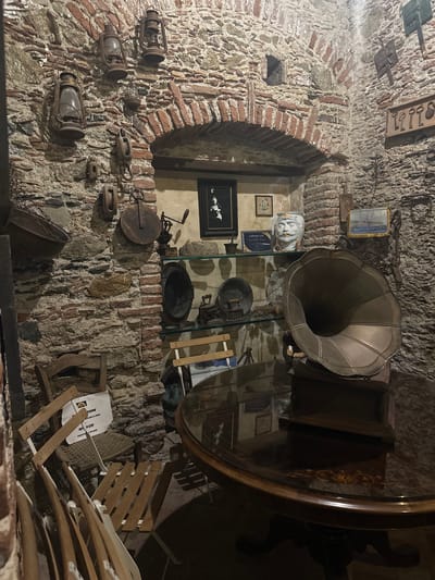The image shows an interior scene, likely a museum or antique shop, possibly located in the city of Rina, Italy. The space is characterized by stone walls with brick accents, reminiscent of an old-world style architecture. Various antique items are displayed, including old lanterns mounted on the wall, pulleys, a gramophone with a large horn, several framed pictures, old cooking pots and tools, and a stone bust. There is also a round table with a mirrored surface, reflecting some of the surrounding objects. A partially visible folding chair is in the lower-right corner, with a sign that includes the words "NO USE" and "ITARE LA SEDIA," suggesting it is not for public seating. A certificate from "Bar Vitelli Savoca" is also visible, adding to the scene's historical context.