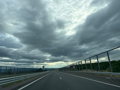 The image depicts a view from a car traveling on a highway. The road is asphalt with white dashed lines marking the lanes. On either side of the road, there are metal guardrails and fences. Beyond the fences, there are trees and hills. The sky is filled with dramatic, dark clouds, suggesting an impending storm or overcast weather. There are two traffic signs visible on the right side of the road, both displaying a red circle with a diagonal line, indicating a prohibition. One sign also has text below it. The location is identified as the city of Chokoba, Bulgaria. There are no people or animals visible in the image. The scene is captured from a moving vehicle, indicated by the slight blur of the surroundings.