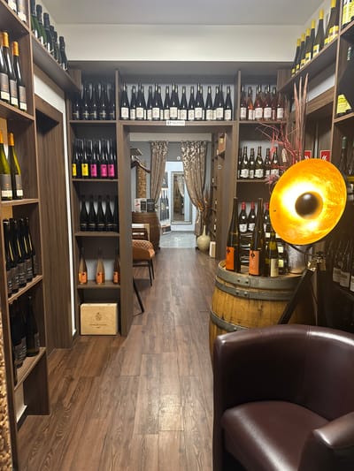 The image shows the interior of a wine shop, potentially located in Grevenmacher, Luxembourg, given the presence of wine bottles. The camera is positioned at floor level, angled upwards. A wooden wine rack filled with bottles of wine dominates the left side of the image. A wooden barrel, serving as a table or display, is situated to the right, with a gold-colored lamp positioned above it. A brown leather chair is also visible. The floor is made of wood planks, running horizontally. In the background, there is an arched doorway leading to another room, with chairs and a mirror visible in the distance.