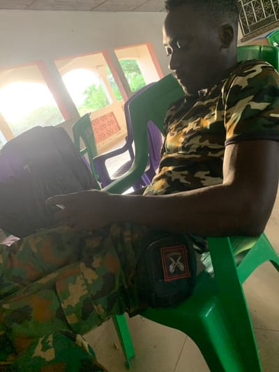 An individual wearing a camouflage uniform is seen in a brightly lit room. They rest their head on a green plastic chair. The person appears to be seated among multiple similar chairs, one of which is purple. A backpack sits nearby. The individual's uniform features a patch with crossed swords on the right sleeve. Outside the window, greenery is visible.