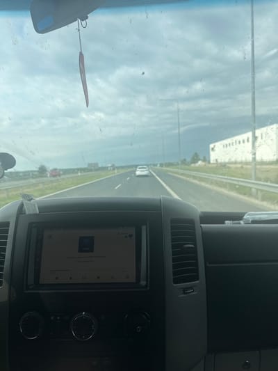 The image shows the interior of a vehicle driving on a highway. The dashboard is visible, with a car stereo displaying music information. A pink air freshener hangs from the rearview mirror. The windshield is dirty, with streaks and smudges. Outside the vehicle, the highway stretches ahead, with other cars visible in the distance. There are streetlights and buildings lining the road. The sky is cloudy and overcast. The location is stated to be Stawiski, Poland, though this cannot be visually confirmed from the image alone.