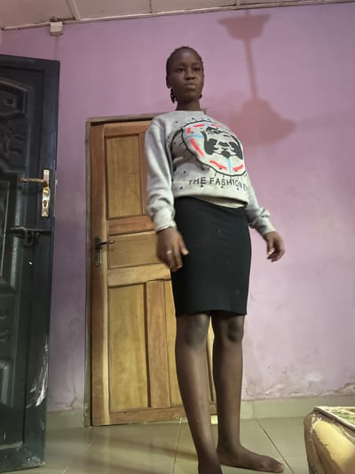A young female stands barefoot indoors in Umuoye, Nigeria. She has dark skin and her hair is styled in braids pulled back. She is wearing a light grey long-sleeved sweatshirt featuring a graphic design of a stylized animal head in black, red, and blue, with "THE FASHION EVE" text printed below it. She is also wearing a plain black fitted knee-length skirt. A ring is visible on her left ring finger. She stands upright, facing the viewer with a neutral expression. The background consists of a pale lavender-pink wall displaying areas of discoloration and wear, particularly near the floor line. Two doors are present: a dark-colored (likely black or dark brown) decorative panel door with a brass-colored handle and a secondary black latch on the left, and a lighter-colored wooden panel door with a dark handle positioned centrally behind the subject. The floor is covered with light-colored tiles. A distinct shadow of a ceiling fixture, possibly a fan or light, is cast on the upper part of the wall behind the subject. A fragment of light-colored upholstered furniture is visible in the bottom right corner of the frame.