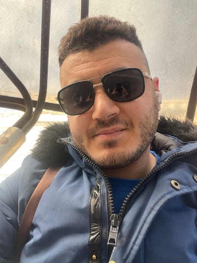 A male individual is depicted in a close-up selfie, situated within an enclosed environment in Zarautz, Spain. The subject, an adult male with short brown hair, a beard, and a mustache, is wearing dark aviator-style sunglasses that reflect the camera phone used to capture the image. A white earbud is visible in his right ear. He is dressed in a dark blue winter jacket featuring a black faux fur collar/hood trim, a prominent silver zipper with a "PAC" branded pull tab, and a shiny black vertical accent strip. A bright blue long-sleeved shirt is visible beneath the jacket, and a brown strap, likely from a bag, crosses his left shoulder. The background is blurred but shows metallic structural elements, including dark bars and a lighter-colored pipe or railing, against a translucent or frosted panel allowing diffuse light. The setting appears to be an interior space, possibly public transport or a sheltered area.