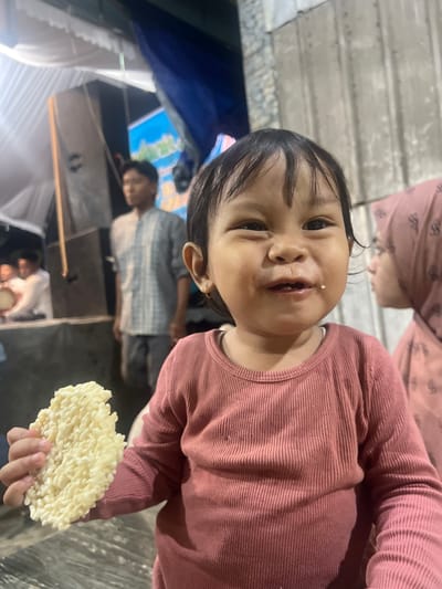 A young Indonesian child in Wedarijaksa, Indonesia, is shown in a close-up portrait, holding a light-colored, textured snack in their left hand and wearing a pink ribbed long-sleeved shirt. The child has dark hair, brown eyes, and appears to be smiling or making a funny face, with a small crumb on their chin. In the background, out of focus, a woman in a pink headscarf is visible to the right, and other individuals are present, along with tents and what appears to be a stage setup. The environment suggests an outdoor gathering or event, possibly a festival or market.
