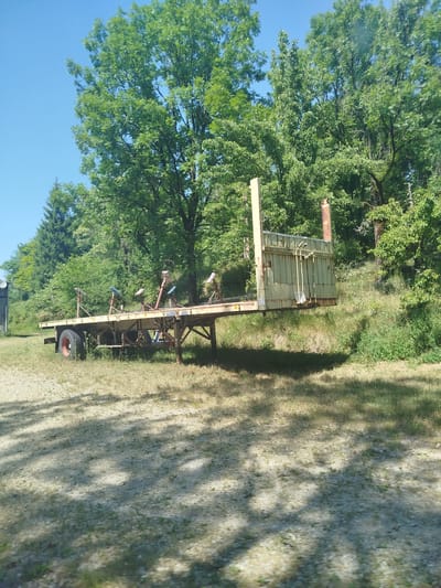 The image shows an empty, weathered, pale green trailer with a yellow vertical section at the rear, resting on a grassy area with scattered gravel and dappled sunlight. The trailer has multiple supports with blue and pink rollers on top, possibly for carrying a boat or similar object. It has one visible wheel with a red rim. The background consists of lush green trees and foliage under a clear blue sky, suggesting an outdoor, possibly rural or park-like setting. The specific location in Charchilla, France, is not discernible from the visual evidence alone; this context would require external information.