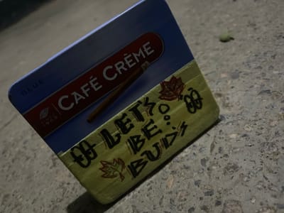 The image displays a close-up, low-angle shot of a rectangular metal tin, likely for tobacco products, resting on a textured, grey concrete or asphalt surface. The tin's upper portion is dark blue with a horizontal red band. White lettering on this band reads "CAFÉ CRÈME," with a small illustration of a cigar directly beneath it. The word "BLUE" is visible on the upper left edge of the tin, and "SINCE 1963" is partially discernible below the "CAFÉ CRÈME" text.
The lower section of the tin features a light green surface, possibly a custom sleeve or decoration. On this green area, a hand-drawn message in black reads "LET'S BE BUDS." This text is flanked by doodles of stylized leaves, resembling cannabis leaves, one outlined in red and another in black, along with other abstract circular marks.
The ground beneath the tin is rough, composed of concrete or asphalt, showing small pebbles and grit. A small, out-of-focus green object is visible on the ground in the blurry background. The lighting is dim, suggesting it is evening or night, with localized illumination highlighting the tin. The setting is outdoors, consistent with a general ground surface in Jama A, Nigeria.