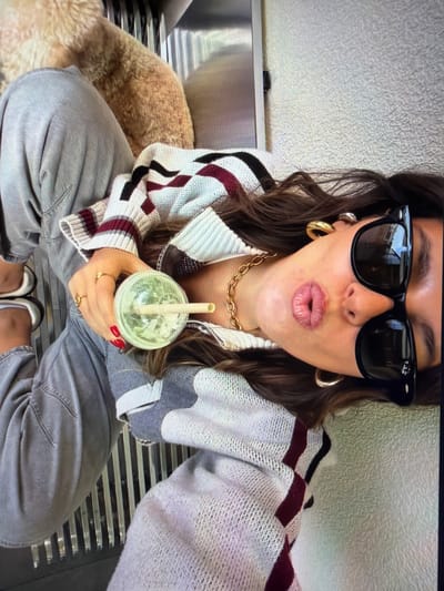 This high-angle selfie captures a person, likely female, sitting indoors on a metal slatted bench in what appears to be Ros Na Rí, Ireland. The subject is seen from the chest up, with her legs and feet also visible. She has dark, wavy hair and is wearing black sunglasses, a thick gold chain necklace, and prominent gold hoop earrings. Her lips are puckered in a playful or kissing expression, enhanced by a light-colored lip gloss.

She is dressed in a zip-up knit sweater or jacket featuring a distinctive pattern of light grey, white, black, and maroon stripes and abstract shapes. Her fingernails are painted a vibrant red, and she wears multiple gold rings on her fingers. In her right hand, she holds a transparent plastic cup containing a green beverage, possibly a smoothie or matcha, with a light-colored straw.

To her left, partially visible, is a fluffy, light brown animal, likely a dog, resting beside her on the bench. She is wearing loose-fitting grey pants and two-toned flat shoes, possibly white and black. The background includes a textured, light-colored wall. The brand name "Ray-Ban" is visible on the temple of her sunglasses. The image conveys a casual and relaxed atmosphere.