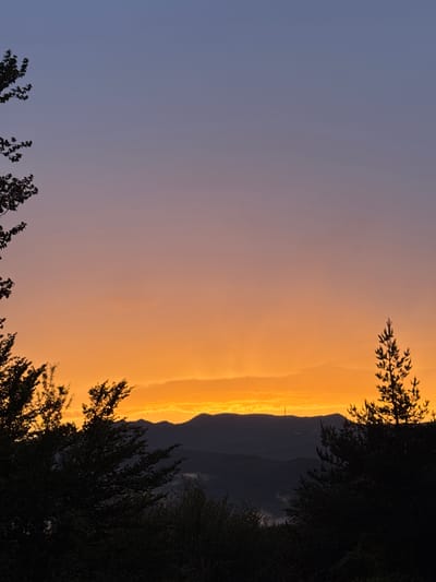 This image captures a sunset over the city of Strazha, Bulgaria. The sky transitions from a deep blue at the top to a vibrant orange and yellow near the horizon. Silhouetted trees frame the scene on the left and right sides. In the background, a range of dark, rolling hills and mountains is visible, with a prominent communication tower standing atop one of the peaks. Wisps of mist or low clouds can be seen in the valleys between the mountains. The overall impression is one of natural beauty and tranquility as day turns to night.