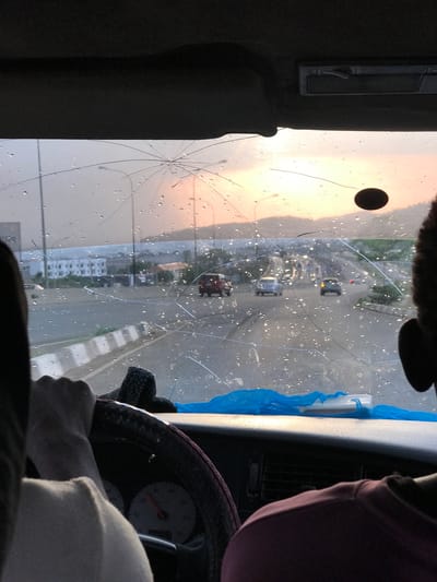 The image is taken from inside a vehicle with a severely cracked windshield. The vehicle appears to be stationary, facing an urban street. Raindrops are visible on the glass. Through the damaged windshield, one can see cars on the road, buildings, streetlights, and a sky with an orange hue, suggesting either sunrise or sunset. A steering wheel is in the foreground, with a person's hands gripping it. A speedometer is partially visible. To the left of the windshield is a dark-colored interior panel of the vehicle with a metallic fixture. To the right is another section of the dashboard, accompanied by a piece of blue fabric. The location is indicated as Jiru, Nigeria.