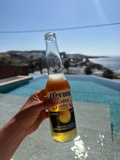 A tanned arm, belonging to a person with manicured fingernails, holds a bottle of Corona Extra beer horizontally. The bottle is partially empty. Behind the bottle, there's a turquoise, shimmering pool with sunlight reflected off its surface. In the blurred background, elements of Mykonos, Greece are visible, including white buildings and hills under a blue sky.