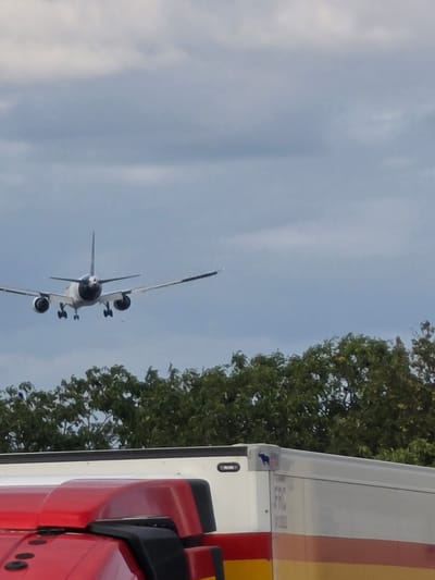 An airplane is in flight above a tree line, with its landing gear extended, suggesting it is preparing to land. Below the airplane, and partially obscuring the tree line, are the roofs of two vehicles, a red truck and a white trailer. The truck is in the foreground and appears to be a transport vehicle. The trailer is behind the truck, and has "FRC" printed on its side. The sky is overcast with light blue and white clouds. The image is taken from a low angle, looking upwards.