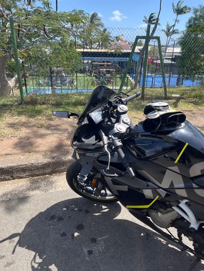 A black CFMOTO motorcycle, with neon yellow accents and large "CFMOTO" lettering on its side fairings, is parked on an asphalt surface adjacent to a concrete curb. A black and white helmet and black gloves rest on the motorcycle's fuel tank. The motorcycle features a dark tinted windshield and aftermarket rearview mirrors. Strong sunlight casts distinct shadows from the motorcycle and nearby trees onto the asphalt.
In the background, behind a chain-link fence, is the "PADEL CLUB DU MONT COFFYN," identified by a blue sign. Inside the fenced perimeter, several individuals are visible near what appear to be blue padel courts. Green support structures and a swimming pool are also partially visible within the club grounds. Lush green foliage, including palm trees and deciduous trees, frames the scene. The sky is clear blue with sparse white clouds. This location is within the city of Nouméa, France.