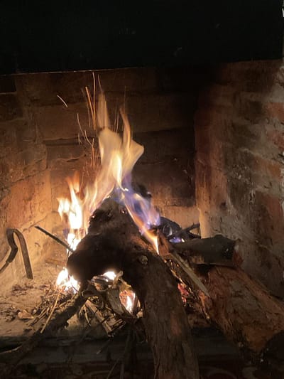 The image shows a close-up view of a fire burning in a brick fireplace. The fireplace appears to be located in El Rodeo, Argentina. The fire is burning with bright orange and yellow flames, with some blue tinges visible. Sparks are flying upwards from the fire. Several logs and branches are burning in the fireplace. The interior of the fireplace is made of brick and appears to be old and weathered. The overall scene conveys a sense of warmth and coziness.