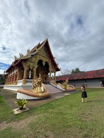 A gilded Thai temple stands beside a green lawn with a person in black shorts and a t-shirt. The temple is intricately decorated and features multiple golden serpent statues. The sky is overcast with white and gray clouds. A building with a red roof is visible in the background.
