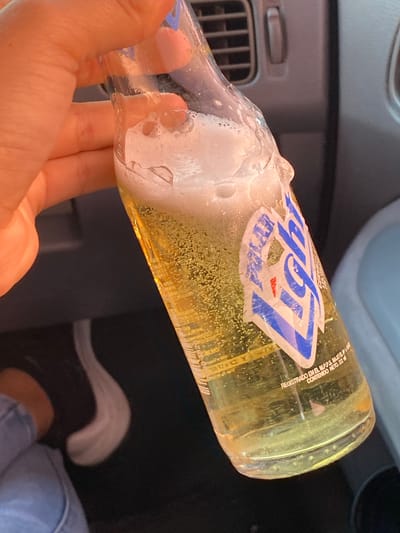 A close-up, eye-level shot captures a person's hand holding a clear glass bottle, likely inside a car in La Cruz del Pastel, Venezuela. The bottle contains a light yellow, effervescent liquid, indicative of a cold beverage such as beer, with significant condensation and foam around its neck and upper body, suggesting it was recently opened or is being consumed.

The subject's hand, with light skin, firmly grips the bottle near its base. The bottle features a partially peeled and damaged blue-and-white label. Prominently visible on the label is the word "Light" in stylized blue font, with "Polar" faintly discernible beneath it, identifying the brand as Polar Light, a Venezuelan beer. Other visible text fragments include "REGISTRADO EN EL MPPS BAJO E..." (Registered with the Ministry of Popular Power for Health under E...) and "CONTENIDO NETO: 0.22..." (Net Content: 0.22...), likely referring to 0.22 liters or 222ml. The word "CERVEZA" (Beer) is also partially visible on the label.

In the background, elements of a car interior are visible, including a dark dashboard and an air vent to the left. The lower right portion of the frame shows a person's leg and foot, wearing dark footwear and light blue jeans, indicating a relaxed posture within the vehicle. The lighting suggests daytime, though no direct sunlight is visible. No emotions are discernible from the partial views of the subjects.