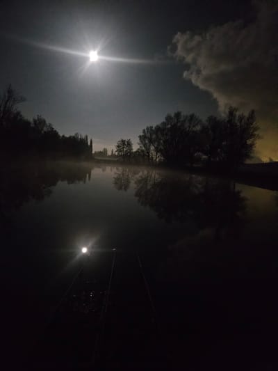 The image shows a night scene, likely in Gavisse, France, based on the prompt. A reflective surface, possibly water, dominates the center, mirroring the sky and silhouetted trees above. A bright light source, possibly the moon, illuminates the left side of the sky, creating a visible beam. Clouds are present on the left side of the sky. Trees are visible on both sides of the reflective surface, appearing dark against the sky. On the right side of the image, another light source is visible, along with what appear to be wires or cables. The overall scene is dark and atmospheric.