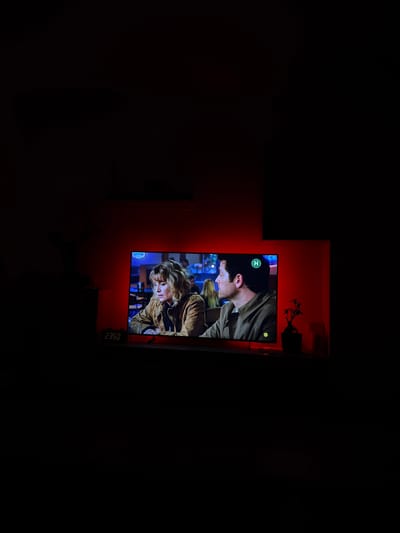 The image depicts a dimly lit room with a television screen displaying a scene from a television show. Two individuals, a woman with blonde hair and a man with dark hair, are visible on the screen. The woman is wearing a brown jacket, and the man is wearing a green jacket. A green circle with an "H" inside is visible on the man's shoulder in the television image. There is a digital clock on the shelf below the television displaying "23:50". A small potted plant is placed to the right of the television. Red ambient lighting emanates from behind the television, illuminating the surrounding wall. The overall environment is dark, suggesting it is nighttime. There is no information in the image to indicate a specific location in the city of Wilhelmshaven, Germany.