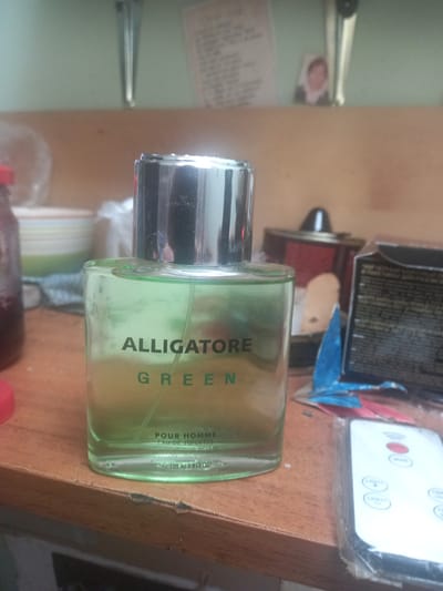 In a room, a bottle of green perfume is displayed on a wooden table. The bottle is rectangular and transparent, with a silver cap. The label on the bottle reads "ALLIGATORE GREEN POUR HOMME EAU DE TOILETTE 100 ml 3.3 FL.OZ". Behind the perfume bottle, there are other objects, including a can, a box, and a remote control. A small framed picture of a person is hanging on the wall in the background. The scene is indoors, with natural lighting. The room appears to be cluttered, with various items scattered around. The floor is not visible in the image. The location of the image is not specified in the provided context.