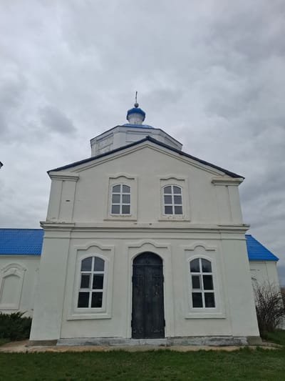 A white, gabled building, likely a church or chapel, stands under an overcast, grey sky. The building's facade is off-white with decorative elements, including pilasters flanking a dark, arched doorway and several windows. It features a bright blue gabled roof and a small blue onion dome with a finial, characteristic of Orthodox Christian architecture. The windows are rectangular with white frames and multiple panes, some elegantly topped with arches. Green grass borders the building on its right side, with some dry, bushy vegetation visible further in the background. The lighting indicates an overcast daytime. There are no people or activities depicted, and no text is visible in the image. This structure is located in Yablonovo, Russia.