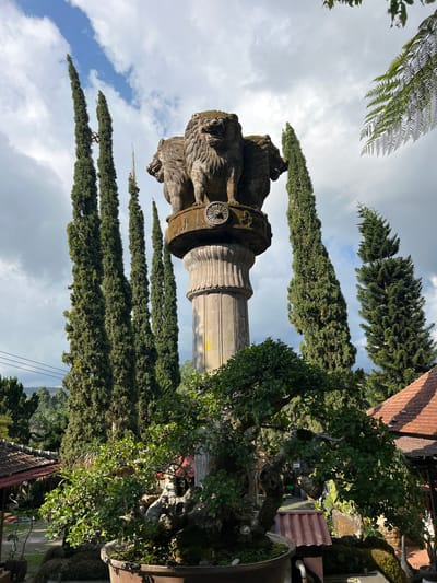 The image shows a stone monument topped with four lion heads, reminiscent of the Ashoka pillar, set against a backdrop of tall cypress trees and a cloudy sky. In the foreground, a large, meticulously pruned bonsai tree dominates the lower half of the frame. The scene appears to be in a garden or park-like setting, with tiled roofs of buildings visible in the middle ground and lush greenery in the distance. The lighting suggests it is daytime, with some areas illuminated by sunlight and others in shadow. The monument and bonsai tree suggest a cultural or aesthetic interest, possibly a tourist attraction or a private collection in Pupuan, Indonesia. There are no people visible in the image.