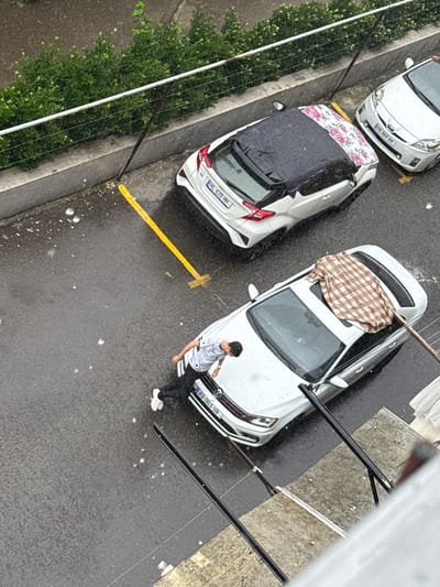 An overhead, high-angle shot captures a man in a patterned shirt and dark pants leaning against a white Volkswagen car. The car has a checkered blanket draped over its roof and a portion of its windshield is visible. The car's license plate reads "AQ-003-UB". In the background, two other white cars are parked on a wet asphalt surface. The car closest to the Volkswagen has a black convertible top with a floral pattern, and its license plate reads "HL-620-HH". The furthest car is a white Toyota Prius with a license plate reading "HV-869-VH". The ground is wet, indicating it is raining, and raindrops are visible in the air. A yellow painted line marks a parking spot. Lush green bushes line a concrete wall behind the parked cars. The scene appears to be in an urban environment, likely a parking area in Tbilisi, Georgia, given the license plate format.