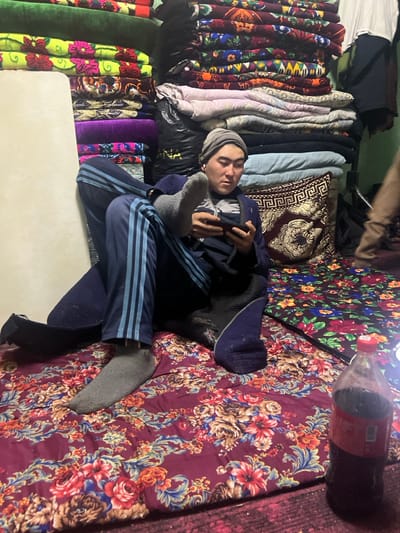 A person lies on a floral-patterned bed, legs crossed, wearing a beanie, blue track suit, and gray socks. They are holding a phone. To the left, stacks of colorful blankets and pillows create a textured backdrop. A large Coca-Cola bottle sits on the top right corner of the bed. The setting appears to be a cozy, possibly cluttered, interior space.