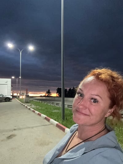 A woman with red curly hair is taking a selfie outdoors at dusk. She is wearing a light blue hooded sweatshirt. Behind her, a street light illuminates a paved area with a white and red striped curb. A car is parked to her left. The sky is dark blue with dramatic clouds, and a faint sunset glow can be seen on the horizon. In the distance, trees and some buildings are visible. The location is likely a roadside, possibly a gas station or rest stop, in the city of Можга, Russia.