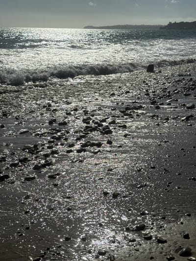 The image depicts a coastal scene on a rocky and wet sandy beach in Malibu, United States. The foreground and midground are dominated by dark, smooth pebbles and rocks scattered across wet sand. Intense sunlight reflects off the wet surfaces, creating numerous bright glints and shimmering effects across the lower two-thirds of the frame. A wave is actively breaking in the midground, moving towards the shore. The ocean surface beyond exhibits significant sun glare, appearing as a broad, shimmering band of light extending towards the distant horizon. A low-lying landmass, indicative of the coastline, is faintly visible in the far background. The sky above the horizon appears overcast or hazy, with a small cloud visible towards the top center. No individuals or distinct man-made structures are visible within the frame.