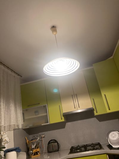 This is a photograph taken from a low angle, looking up at a kitchen ceiling and cabinets. A light fixture with a white, concentric-ringed shade hangs from the ceiling. The kitchen cabinets are a mix of lime green and cream colors with silver handles. To the left, a sheer white curtain hangs. Below the cabinets, a gray backsplash is visible, with electrical outlets and a charging cable. A kitchen stove with two burners is to the right, next to a white colander. On the counter to the left, a wooden utensil holder, a white orchid plant, and a blue cloth are present. A small appliance with a red and gray casing is to the right of the outlets. The scene appears to be an indoor residential kitchen. The location is Jihlava, Czechia.