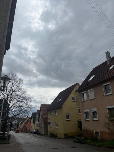 The scene captures a residential street in Hardthausen am Kocher, Germany, under an overcast, heavily clouded sky. The asphalt road features white markings and gently curves into the distance. Along the right side, a row of two-story residential buildings with brown terracotta-tiled pitched roofs extends down the street. The closest building has a pale peach-colored facade with multiple windows, some fitted with partially open white roller shutters, and two skylights on its roof. Adjacent to it stands a building with a yellow facade, also featuring windows and roof skylights. A blue circular speed limit sign indicating "30" is mounted on a pole near the yellow building. Overhead power lines are visible across the upper right sky. On the left, a bare deciduous tree is situated next to an ornate black streetlamp. A dark-colored building with a textured, light-colored facade is partially visible on the far left. A red compact car is seen driving away in the mid-ground, and at least two other dark-colored vehicles are parked along the street. No individuals are present in the observable area.