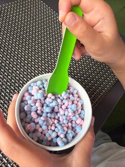 The image shows a close-up of a white bowl filled with small, round, pastel-colored pieces of what appears to be ice cream or mochi. The pieces are a mix of light blue and pink, and they are dusted with a light coating of frost. A bright green plastic spoon is partially submerged in the bowl, held by a person's hands. The bowl sits on a textured surface, possibly a placemat, with a pattern of woven brown and white threads. The background is blurred, suggesting an outdoor or casual setting. The person's hands, one holding the bowl and the other holding the spoon, are visible. The lighting is bright, casting soft shadows. There is no information within the image to indicate a specific location in La Vila Joiosa, Spain.
