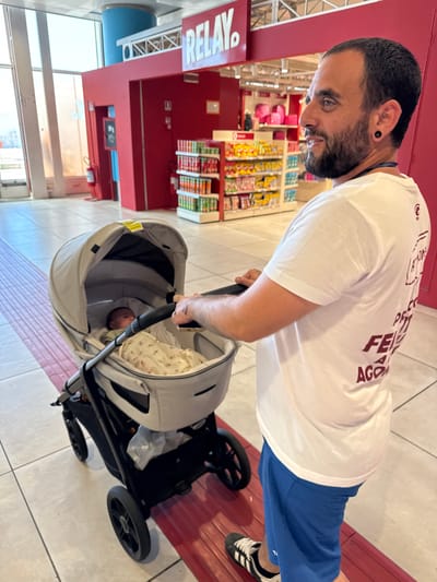 A man with a beard and a white t-shirt is pushing a baby stroller in what appears to be the inside of an establishment in Cinisi, Italy, as a "RELAY" store sign is visible. The man is wearing blue shorts and black and white sneakers. A baby is sleeping in the stroller. There are various products on shelves in the background.