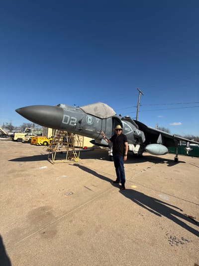 A man in a black shirt, jeans, and brown shoes stands next to a gray fighter jet. The jet has the number "02" painted on its side. A yellow utility vehicle is parked near the jet, with a metal scaffolding adjacent to the jet's body. A shadow stretches from the man along the concrete ground. The sky is clear blue, and a utility pole is visible in the background.