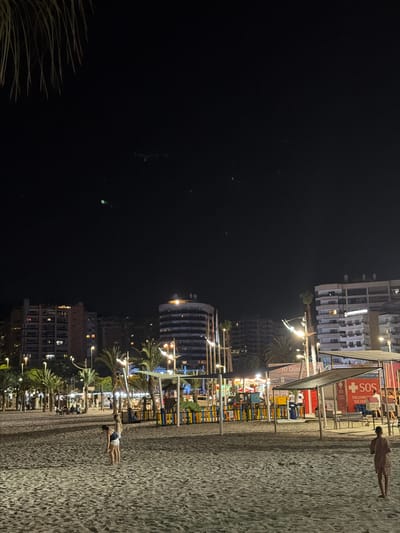 The image depicts a beach scene at night in La Cala, Spain. In the foreground, two young females are walking on the sandy beach, and in the mid-ground, a person is standing on the sand facing away from the camera. The background features illuminated buildings, palm trees, and a playground area with colorful fencing. There are also several lights visible, including streetlights and lights from the buildings. A red and white structure with the word "SOS" is also visible, suggesting a lifeguard or rescue station. The sky is dark and clear.