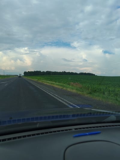 An eye-level medium shot captures a view from inside a vehicle traveling on an asphalt road, presumably in the vicinity of Аздзелінскі сельскі Савет, Belarus. The road stretches ahead, marked by white lines and some damp patches, suggesting recent rain. To the right of the road, a vast, vibrant green cornfield dominates the landscape, extending to a dense line of dark green trees on the horizon. The sky above is filled with a dramatic expanse of grey and white clouds, with patches of pale blue sky peeking through, indicating overcast conditions with some breaks in the cloud cover. A white truck and a dark-colored car are visible on the road in the distance, indicating traffic. The foreground of the image shows the interior of the vehicle, including the dashboard with a blue pen lying on it and a blue patterned trim above the dashboard. The overall scene depicts a rural highway landscape under a dynamic, cloudy sky.